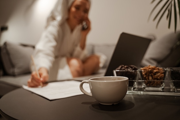 Close-up concept. A business woman in a white robe and a towel uses a laptop and drinks tea or coffee. She sits on the sofa and writes documents. Evening relaxation.