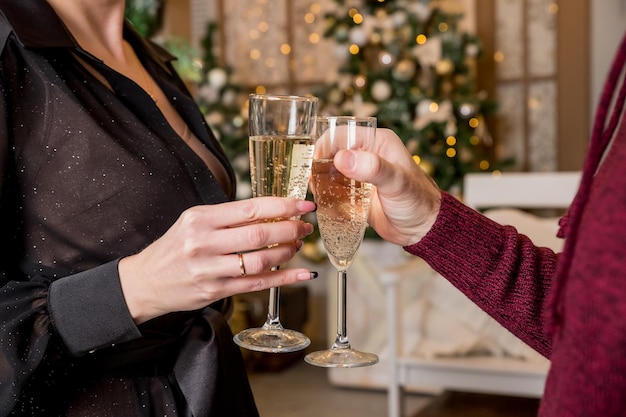 Close up of couple hands holding glasses of champagne over Christmas tree with lightsCouple celebrate Christmas winter holidays at home together wish luck and joy