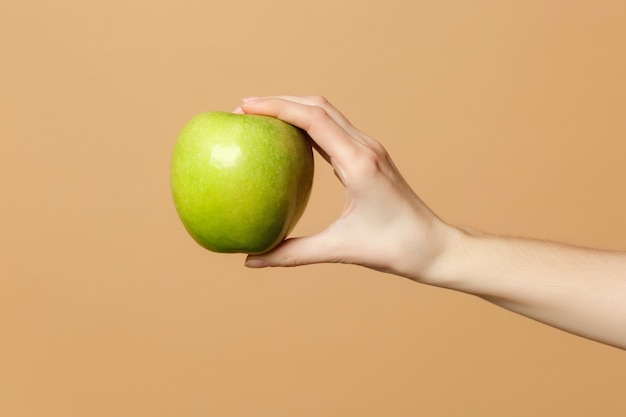 Close up cropped of female holds in hand fresh ripe green apple fruit isolated on beige pastel wall