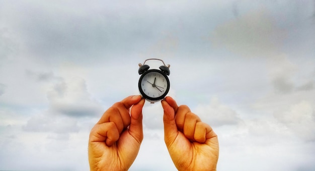 Photo close-up of cropped hands holding alarm clock against cloudy sky