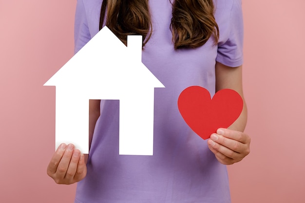 Close up cropped young female wears t-shirt demonstrates small white paper house and little red heart, isolated on pink background in studio. Concept of loving, protecting, cozy home, Valentine's day