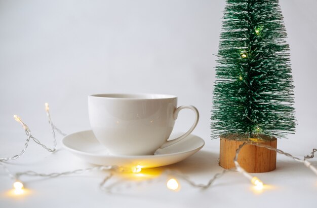 Close-up of a cup with a saucer and a small artificial Christmas tree on a white background with a place for text