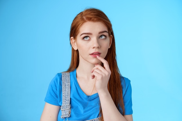 Close-up curious dreamy creative redhead female working project, student thinking essay, touch lip intrigued, look up thoughtful, pondering choice, make assumptions, stand blue background