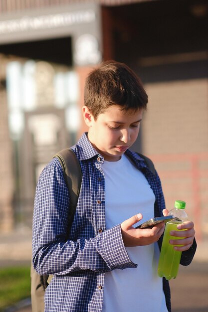 Photo a close up of cute student boy with backpack holds a mobile phone and bottle of water outdoor