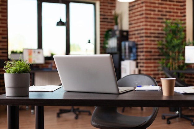 Close up of desk with laptop, coffee cup and plant pot in empty startup office. Nobody in global business space with computer, drink and modern decorations on wooden furniture.