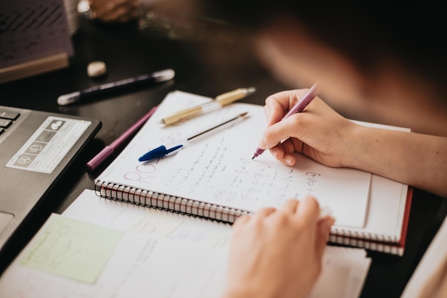 Close up detail image of a young woman studying and working on his home desk doing homework during university preparing for exam with textbook and taking notes selective focus on the pencil