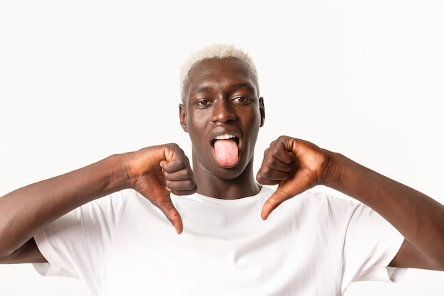 Close-up of disappointed african-american blond male showing thumbs-down and stick tongue from disappointment, dislike and judging something bad