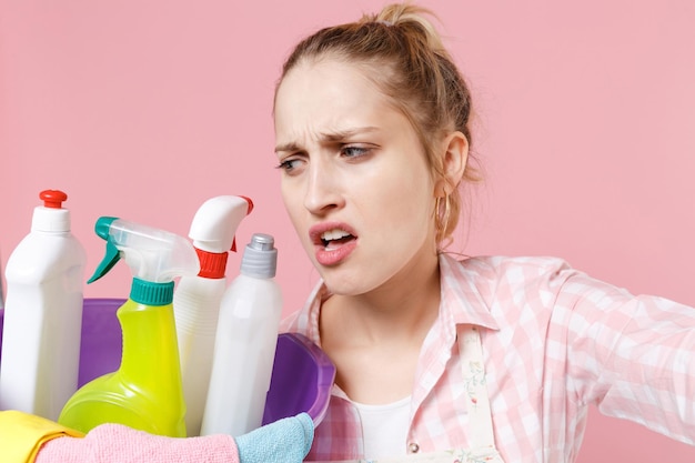 Close up dissatisfied woman housewife in apron hold basin with detergent bottles washing cleansers doing housework isolated on pink background. Housekeeping concept. Doing selfie shot on mobile phone.