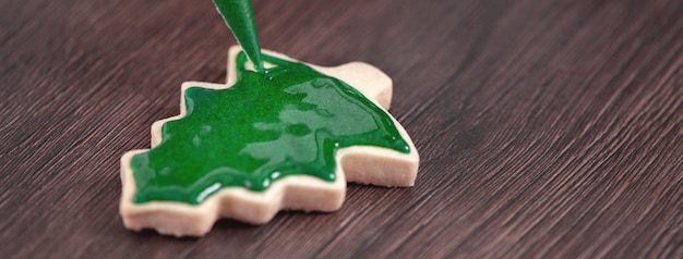 Close up of drawing gingerbread Christmas tree sugar cookie on wooden table background with green icing, concept of holiday celebration.