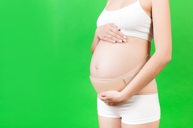 Close up of elastic maternity band dressed on pregnant woman in underwear to reduce backache at green background with copy space. Orthopedic abdominal support belt concept.