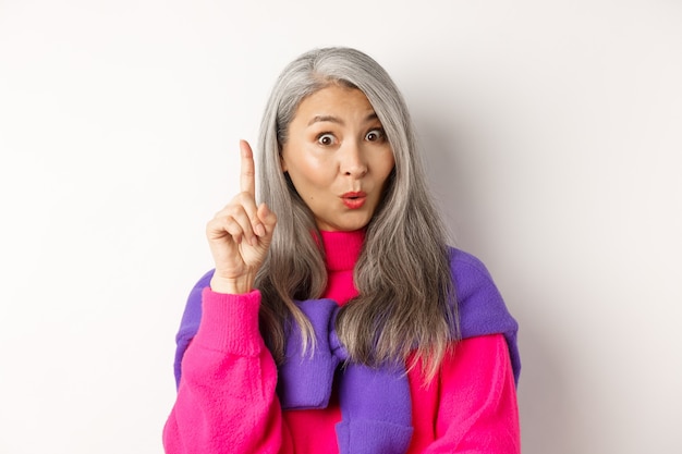 Close up of elderly asian woman in stylish hipster clothes, raising finger and saying idea, have suggestion, standing over white background.