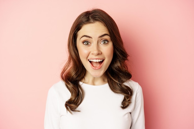 Close up of enthusiastic brunette girl with makeup smiling and looking happy at camera posing agains...