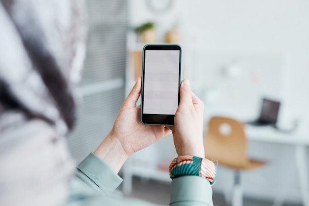 Close up of ethnic young woman holding smartphone in office focus on blank white screen, copy space