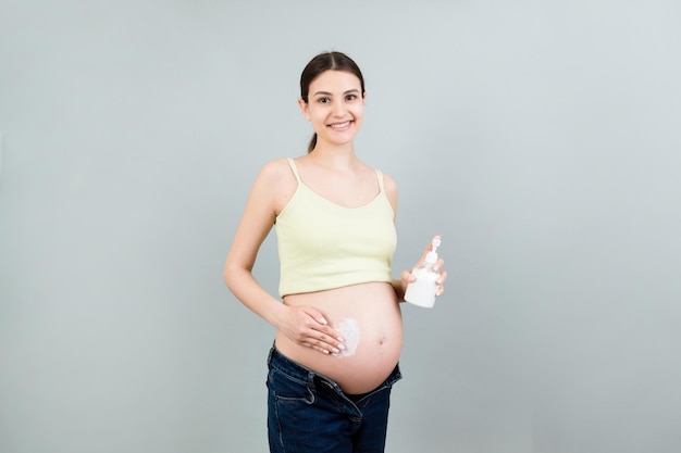 Close up of expecting mother in unzipped jeans applying cream on her pregnant belly at colorful background with copy space Skin care concept