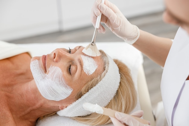 Close up face peeling mask, spa beauty treatment, skincare. Senior woman getting facial care by beautician at spa salon.