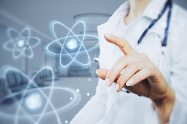 Close up of female doctor hand pointing at abstract glowing atom hologram on blurry hospital or clinic interior background Nuclear medicine energy and medical research concept