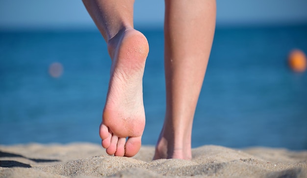 Close up of female feet walking barefoot on white grainy sand of golden beach on blue ocean water background