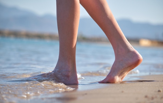 Close up of female feet walking barefoot on white grainy sand of golden beach on blue ocean water background