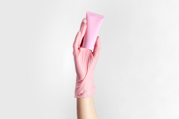 Close-up of female hand wearing glove, holding cosmetic tube of pastel coral pink color, isolated on white studio background.