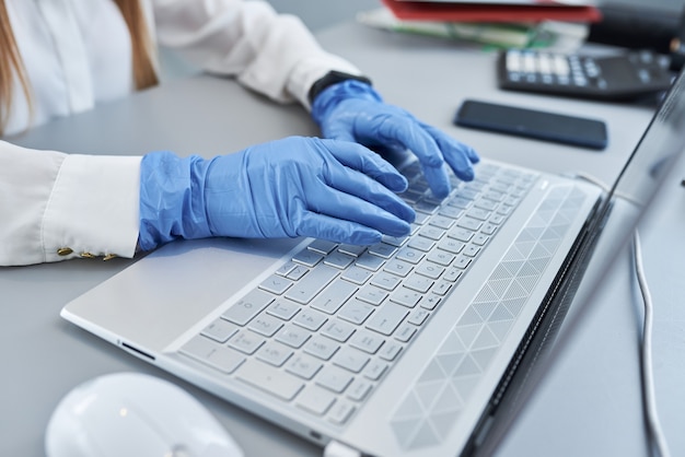 Close up of female hands in blue surgical gloves on laptop keyboard.