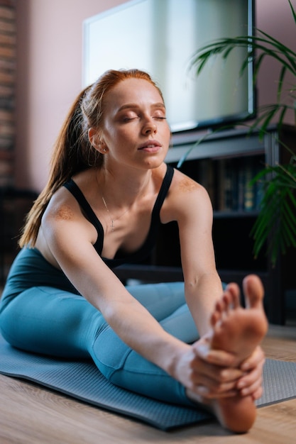 Close-up of flexible redhead young woman working out, doing stretching exercise on yoga mat while watching fitness video online on laptop. Concept of sports training red-haired lady during quarantine.