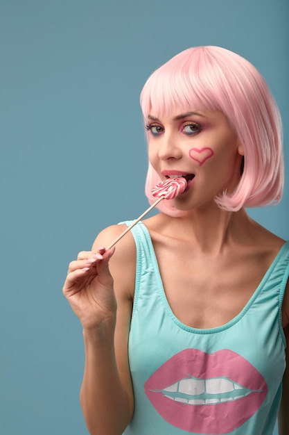 Close up of a flirtatious female model on blue background wearing a pink wig holding different kinds of candies Pretty glamorous woman pink hair charm sweets lifestyle enjoyment sugar