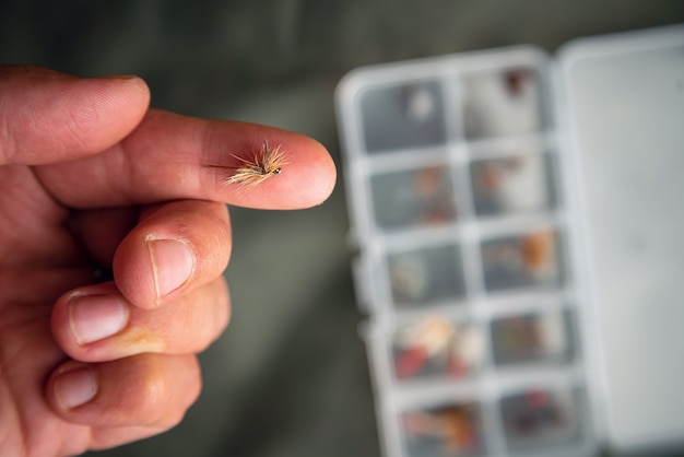 Close up of fly fishing fly on the finger next to box with tied flies. Fly fishing equipment still life.