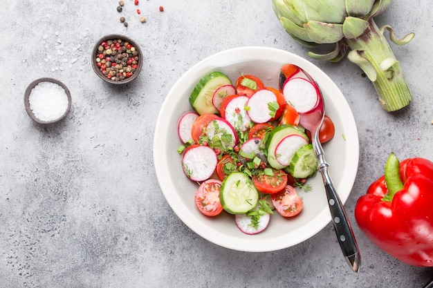 Close-up of fresh healthy salad in a bowl made of tomatoes, cucumber, radish and herbs, with raw artichoke and seasonings, good for diet or detox, grey rustic stone background