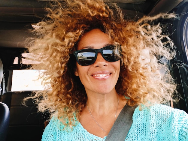 Close up front portrait of young adult woman with lot of curly blonde long hair sitting inside the car as a passenger during travel Pretty woman smiling and having fun with wind and hairstyle