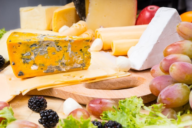 Photo close up of gourmet cheese board featuring bounty different of cheeses and garnished with fresh fruit, served on wooden cutting board