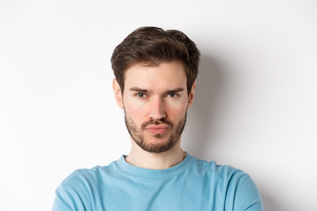 Close-up of grumpy young man grimacing, sulking and making offended face, standing upset over white background.