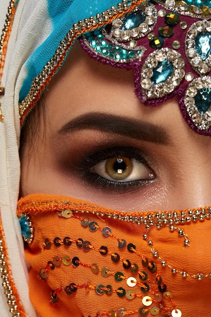 Close-up half face photo of a pretty young woman with professional make-up wearing an elegant colorful hijab decorated with sequins and jewelry. She is posing at the studio on a dark background. Human