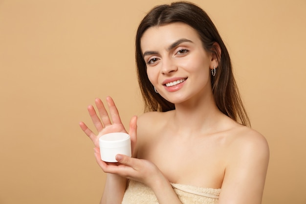 Close up half naked woman with perfect skin nude make up applying facial cream isolated on beige pastel wall