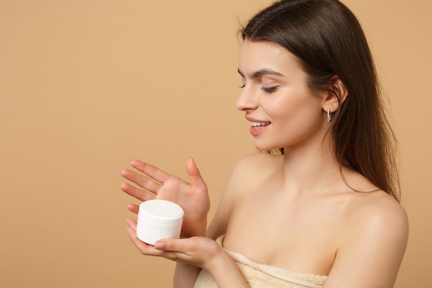 Close up half naked woman with perfect skin nude make up applying facial cream isolated on beige pastel wall