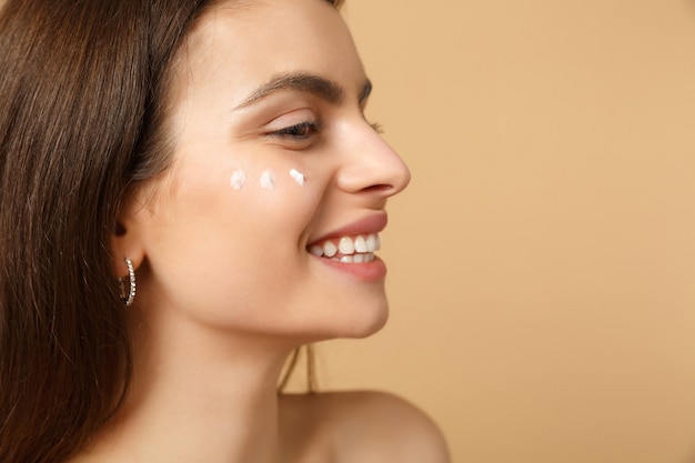 Close up half naked woman with perfect skin nude make up applying facial cream isolated on beige pastel wall