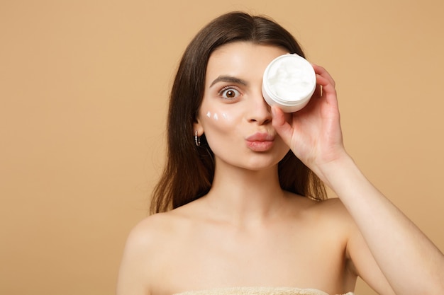 Close up half naked woman with perfect skin nude make up applying facial cream isolated on beige pastel wall