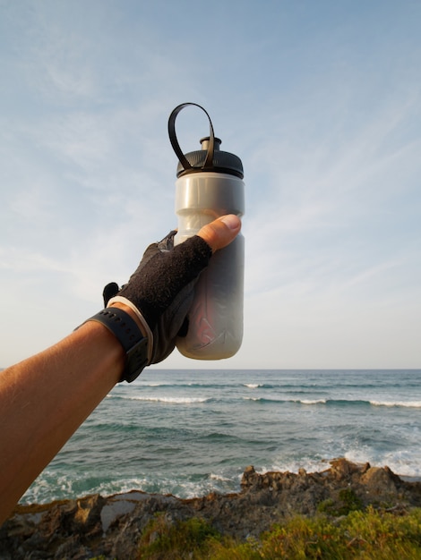 Close-up. Hand of a cyclist in sport glove holds a sports bottle in front of the ocean.