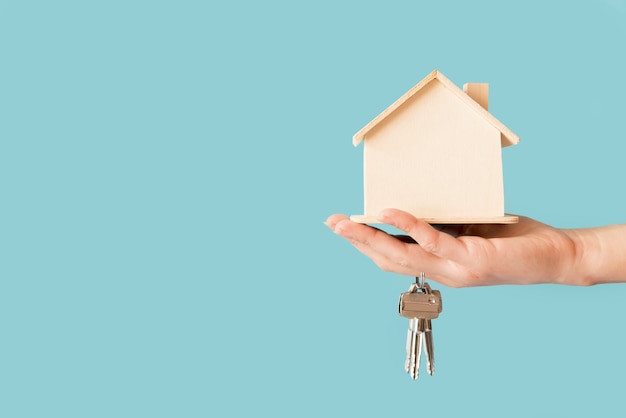 Close-up of hand holding keys and wooden house model against blue background