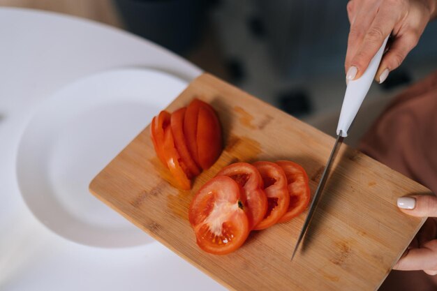 Close-up of hand of unrecognizable woman cutting fresh tomato using kitchen knife on wooden cutting board. Young woman cutting fresh organic tomato with a knife for vegetable salad.