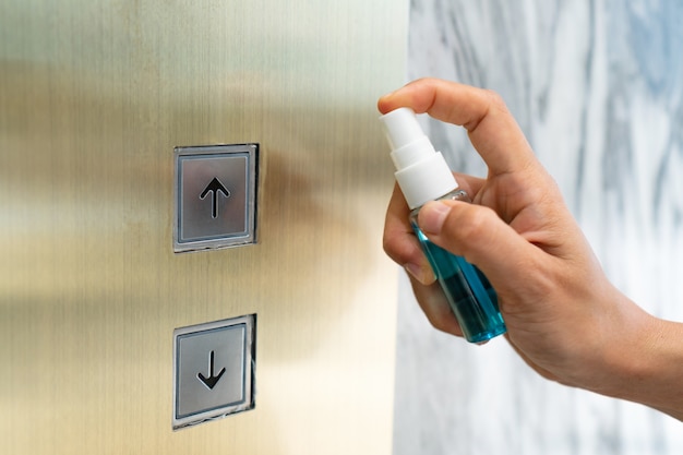 Close up hand of woman disinfecting lift push button by spraying alcohol from a bottle. Protection against infectious virus, bacteria and germs, Coronavirus/ Covid-19, health care concept.