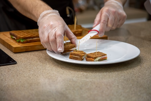 close up of hands of a cook man in rubber gloves put on a white plate snacks grilled sandwiches. close up, soft focus, background is kitchen in blur