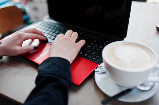 Close up hands of girl working with red laptop with cup of cappuccino.