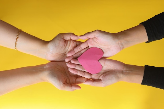 Close up hands of two people holding a red heart shape on yellow background. 