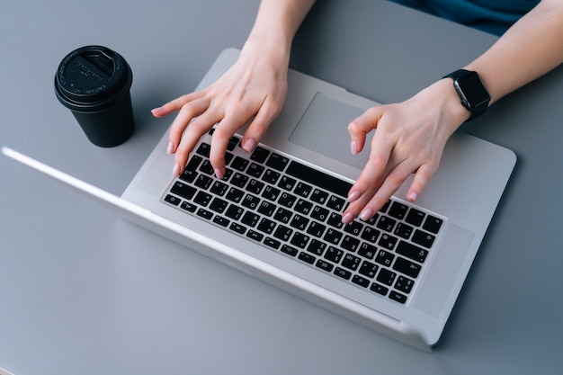 Close-up of hands of unrecognizable business woman typing on laptop keyboard sitting at desk on background of window in office room, selective focus, coffee cup.