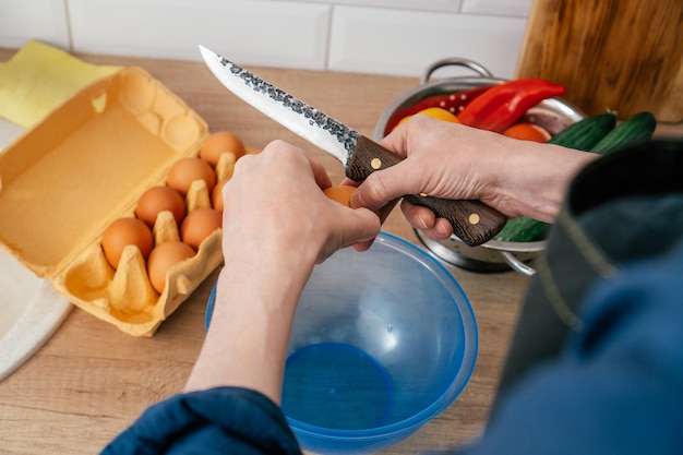 Close up of hands of unrecognizable man breaking egg with knife into blue plastic round bowl near steel colander with yellow bell pepper and wooden cutting board Cooking healthy food diet