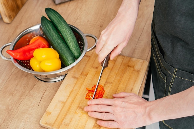 Close up of hands of unrecognizable man chop red bell pepper with knife on wooden cutting board near steel colander