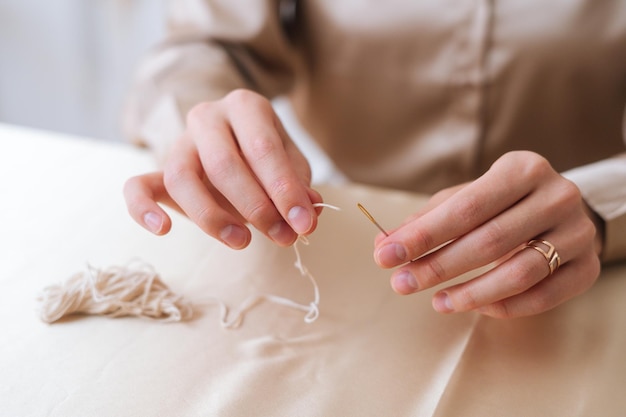 Close-up hands of unrecognizable woman putting thread through eye of needle sitting at table. Closeup of female artisan putting wick through needle. Process of making handmade natural candle