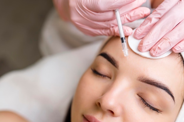 Close up of hands of young cosmetologist injecting botox in female face She is standing and smiling The woman is closed her eyes with relaxation