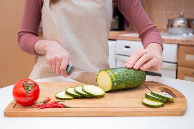 Close-up Hands of a young woman cut with a knife into slices or slices of young zucchini cucumber on a wooden cutting board. Preparation of ingredients and vegetables before cooking and for grilling.