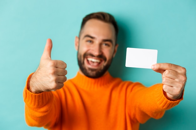 Close-up of handsome caucasian man going on shopping, showing credit card and thumb up in approval, standing over turquoise background.